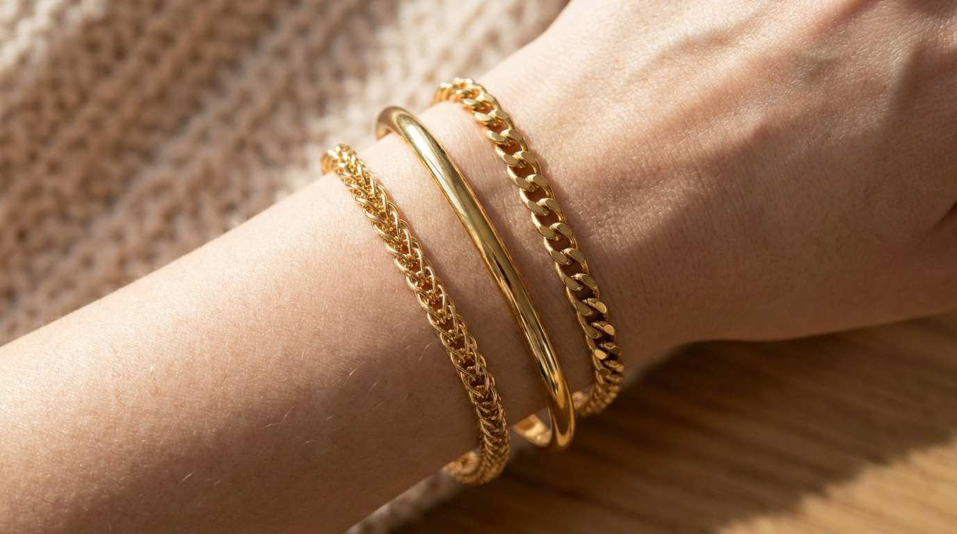 Poignet portant trois bracelets en plaqué or de styles différents sur un fond en maille douce.